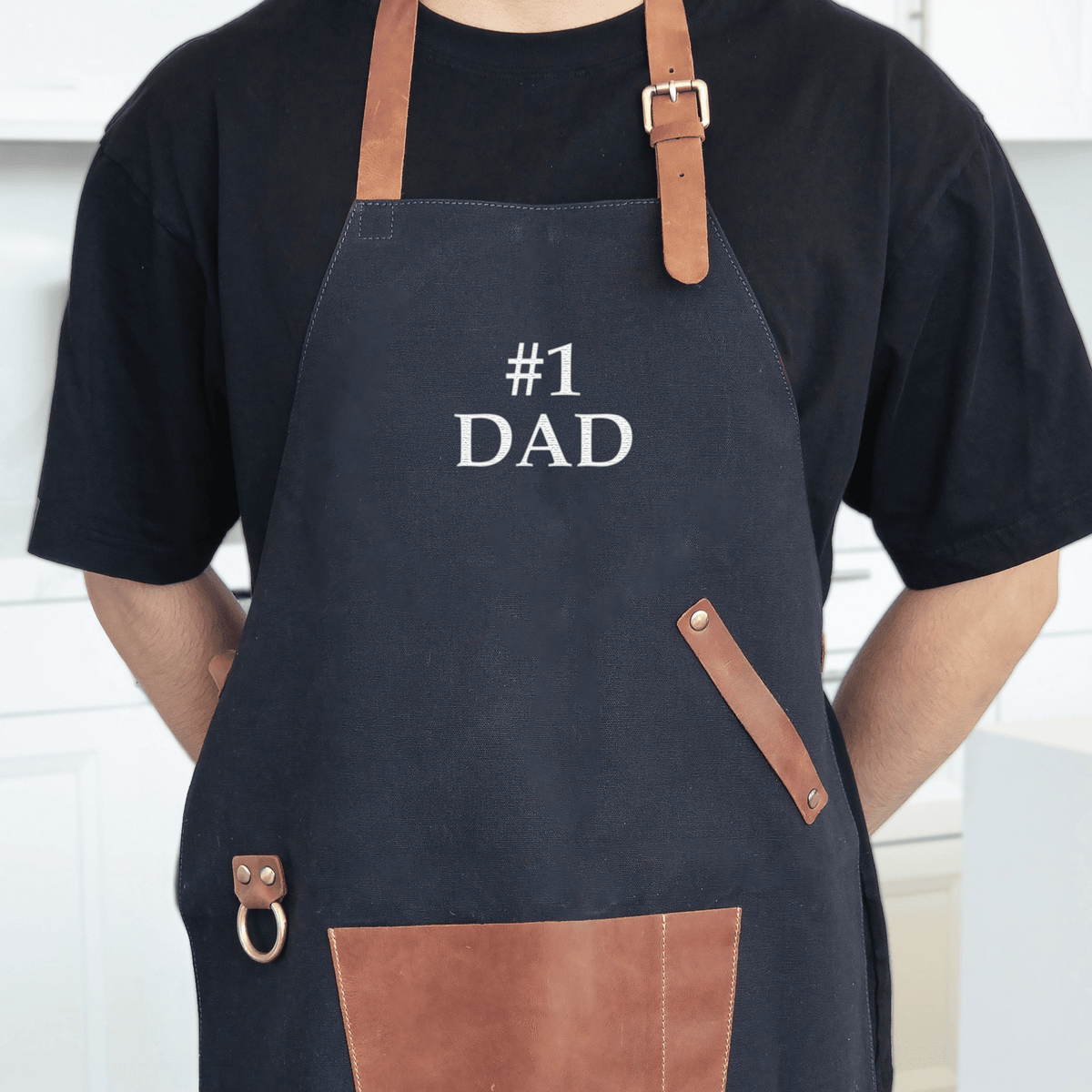 A person wearing a dark canvas apron with leather straps and pocket details embroidered with "#1 DAD" across the chest.