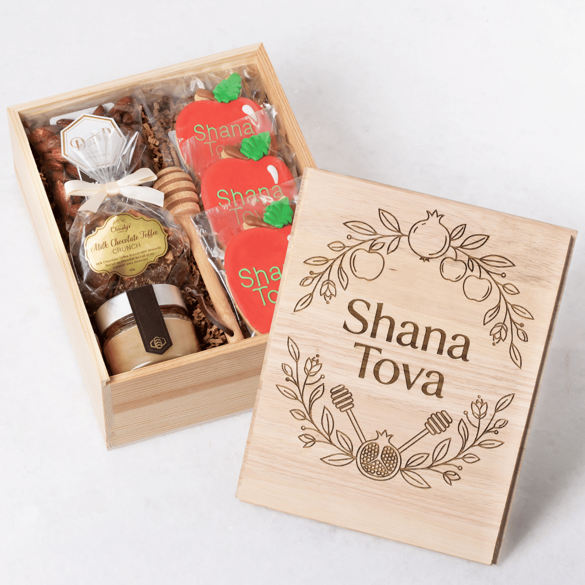 A wooden gift box featuring an engraved "Shana Tova" floral design, displayed next to its contents including honey, a wooden honey dipper, chocolate toffee crunch, and apple-shaped cookies.