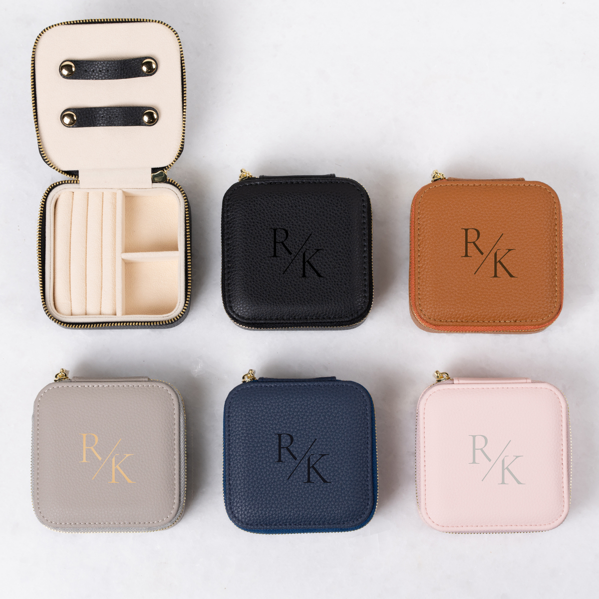 A grid of six monogrammed travel jewelry cases in various colors, including one open case showing ring slots and necklace straps alongside closed black, tan, grey, navy, and pink options.