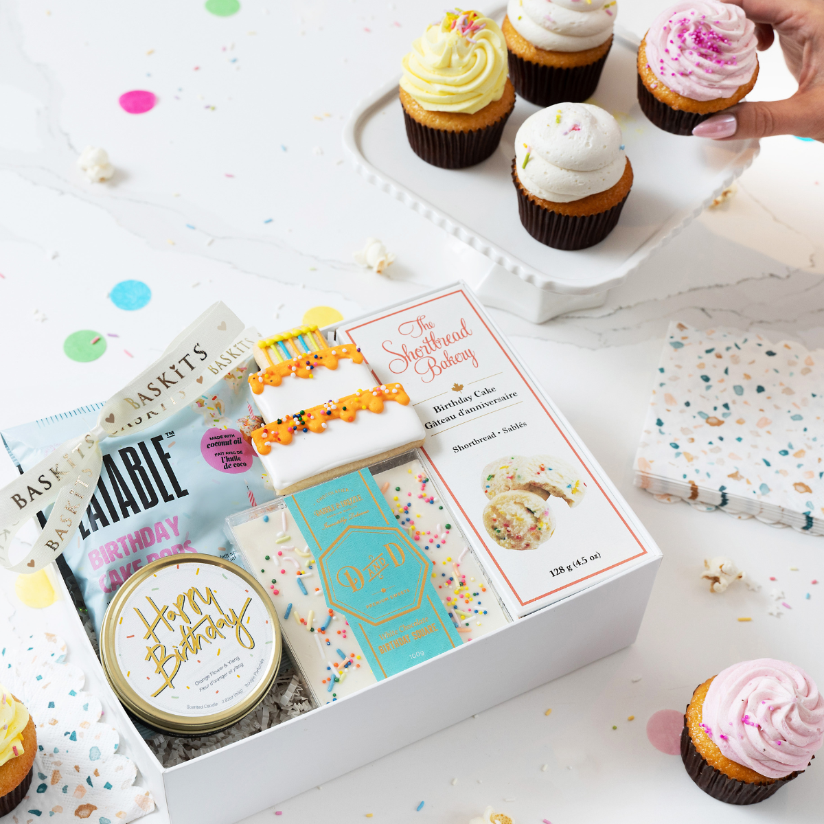 A birthday-themed gift box packed with festive sweet treats and a candle, styled alongside colorful cupcakes.