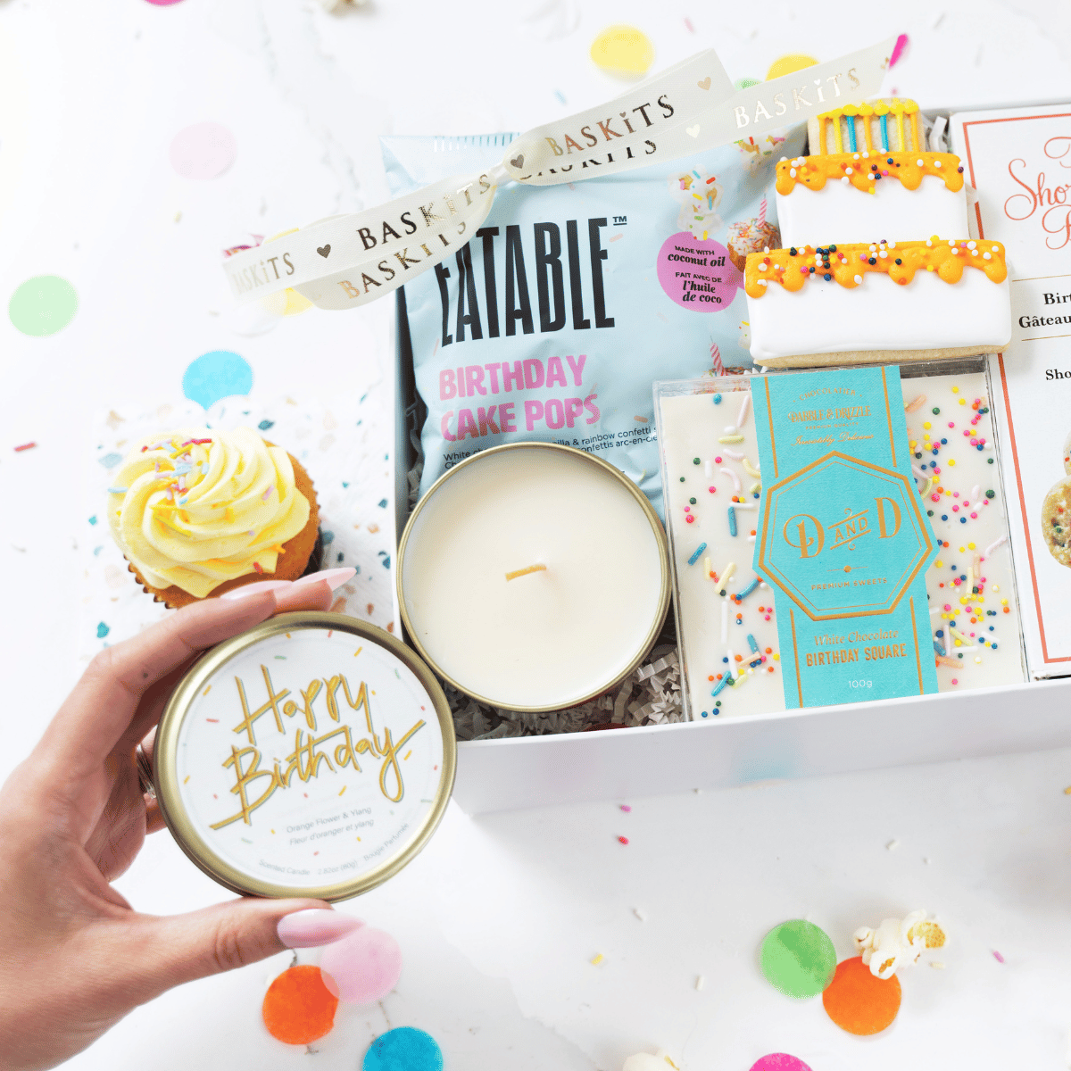 A hand holding the lid of a "Happy Birthday" tin candle next to a opened birthday gift box filled with Eatable Birthday Cake Pops, a Dabble & Drizzle sprinkles chocolate bar, and a decorated cake cookie.
