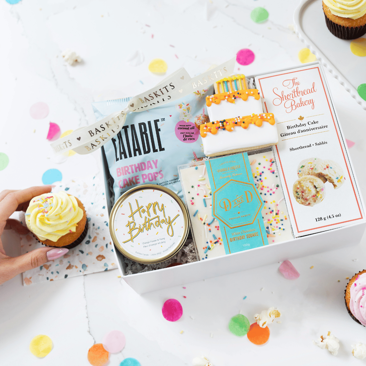 A hand holding the lid of a "Happy Birthday" tin candle next to a opened birthday gift box filled with Eatable birthday cake pops, a Dabble & Drizzle sprinkles chocolate bar, and a decorated cake cookie.