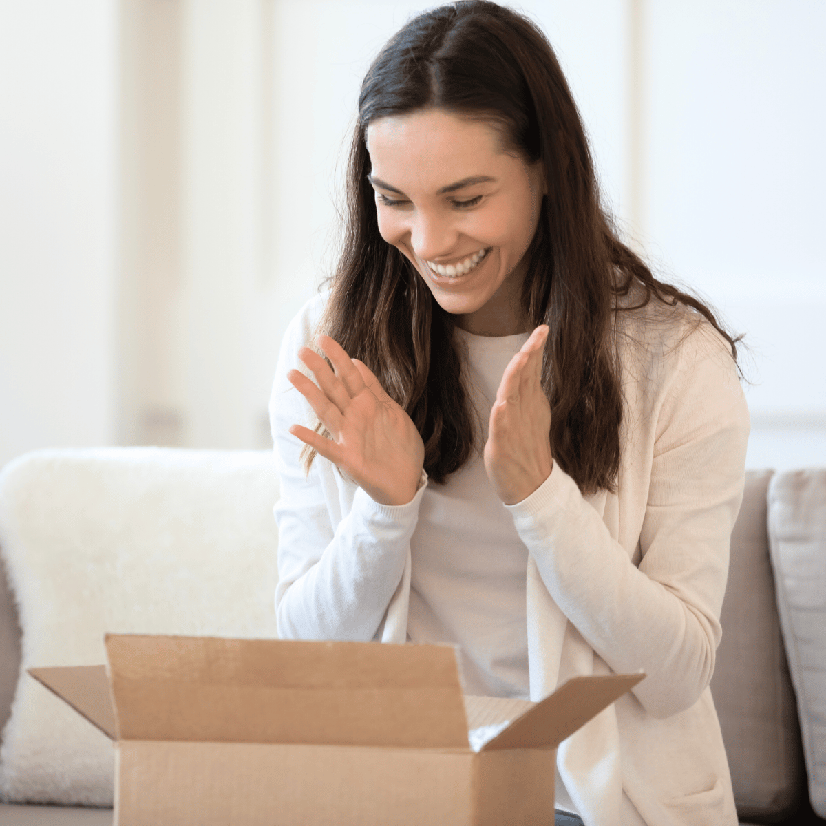 A happy woman sitting on a sofa and clapping her hands with delight as she unboxes an open cardboard package