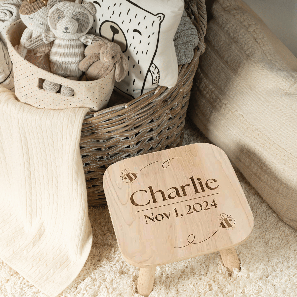 A "Step Stool - Bees" personalized wooden toddler stool engraved with the name "Charlie," the date "Nov 1, 2024," and bee illustrations, set beside a basket filled with plush toys.