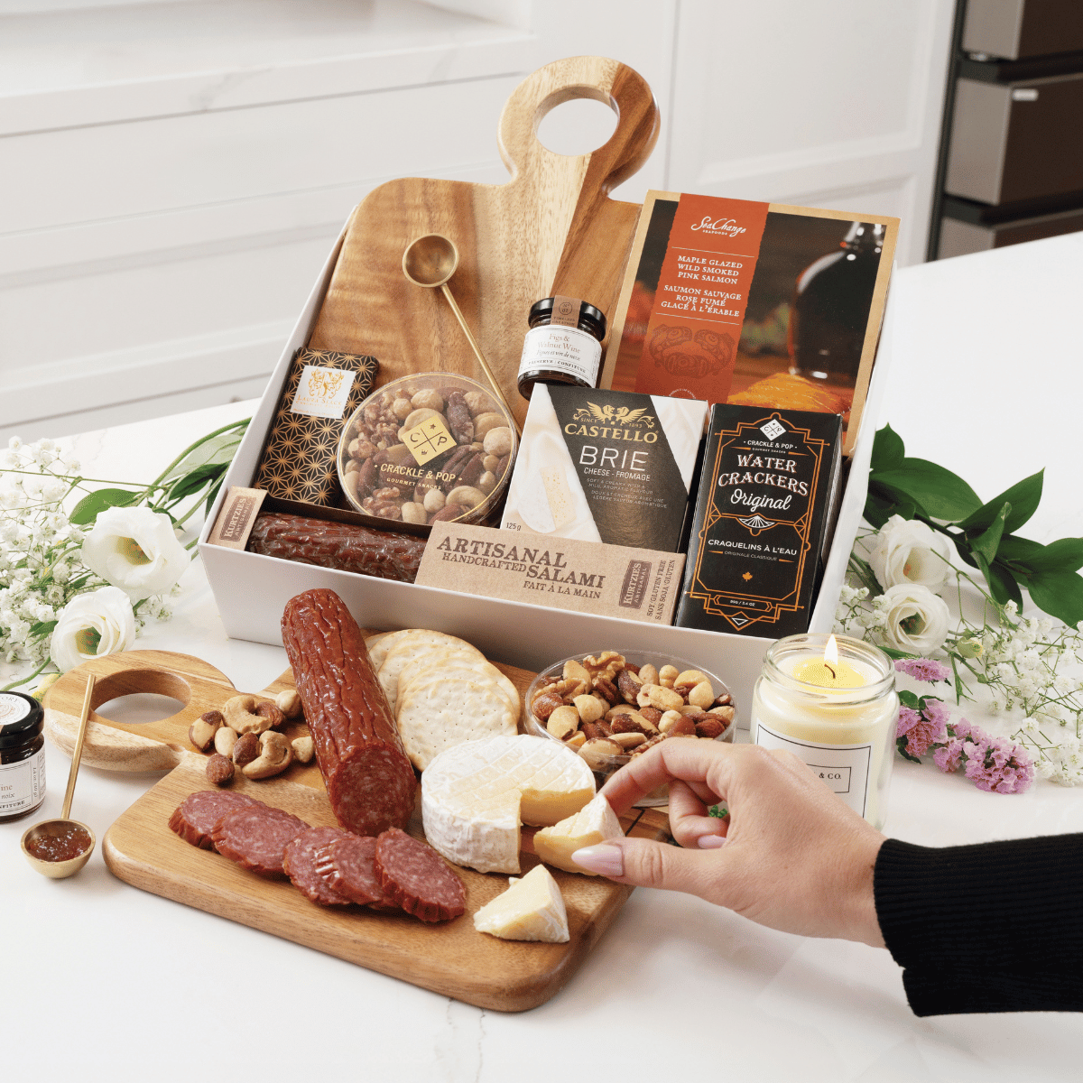 A hand taking a wedge of brie cheese from a charcuterie board with sliced salami and nuts, displayed in front of the Not Too Sweet gift box filled with artisan snacks and a wooden cutting board.
