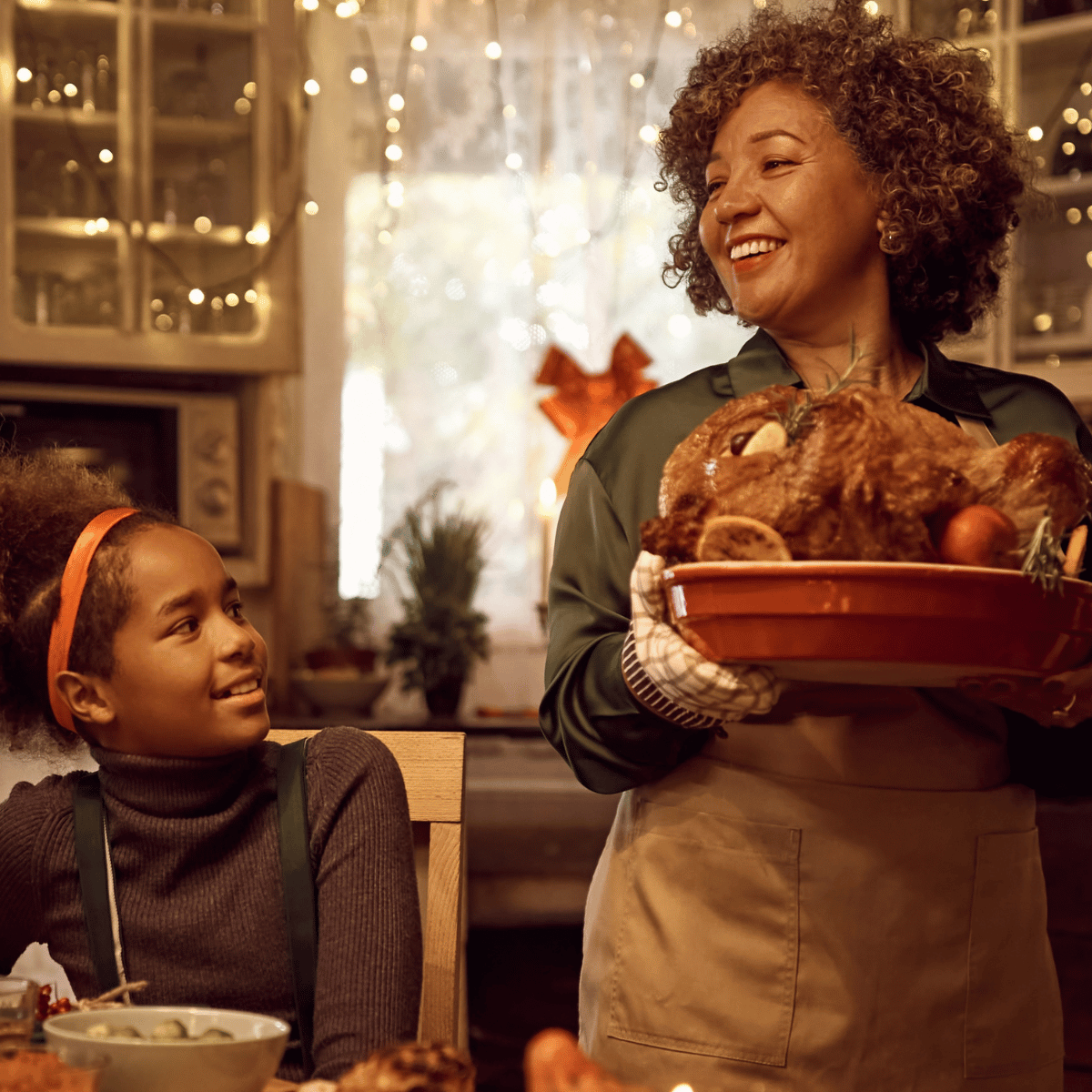 A smiling woman presenting a freshly roasted turkey on a serving platter to a young girl seated at a festive, warmly lit holiday dinner table.