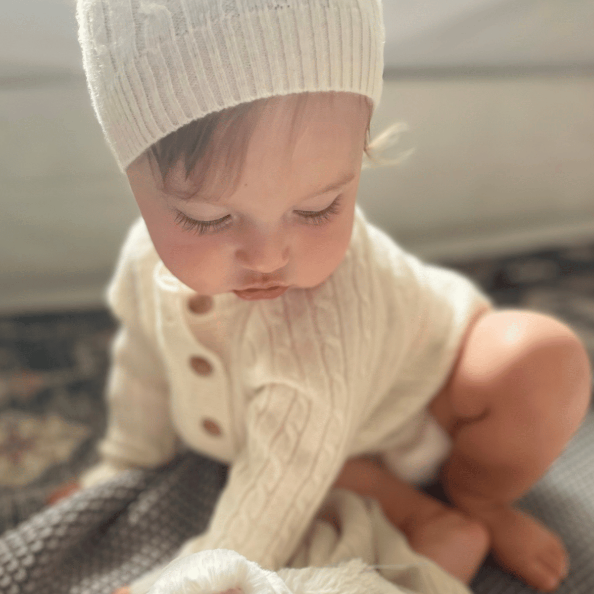 A baby sitting on the floor wearing the Baby Cashmere Set in beige, including a cable-knit sweater cardigan and a matching ribbed beanie.