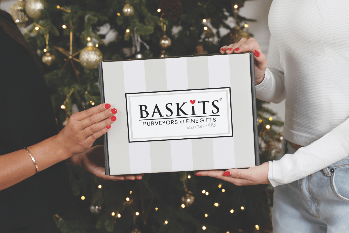 Two people holding a striped gift bag featuring the Baskits logo in front of a warm, twinkling Christmas tree.