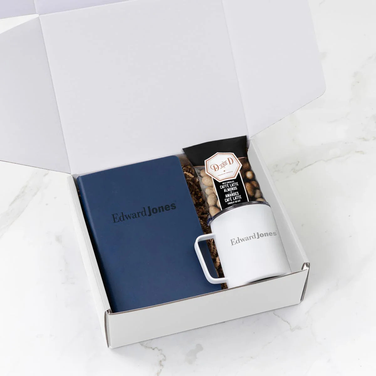 An open white gift box containing custom Edward Jones branded items, including a blue hardbound notebook, a white mug, and a package of Dabble & Drizzle caffe latte almonds.