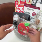 Hands unpacking an 11-piece Shana Tova gift basket, displaying a traditional, Hand-Painted Shana Tova Apple-Shaped Cookie and an array of festive indulgences.
