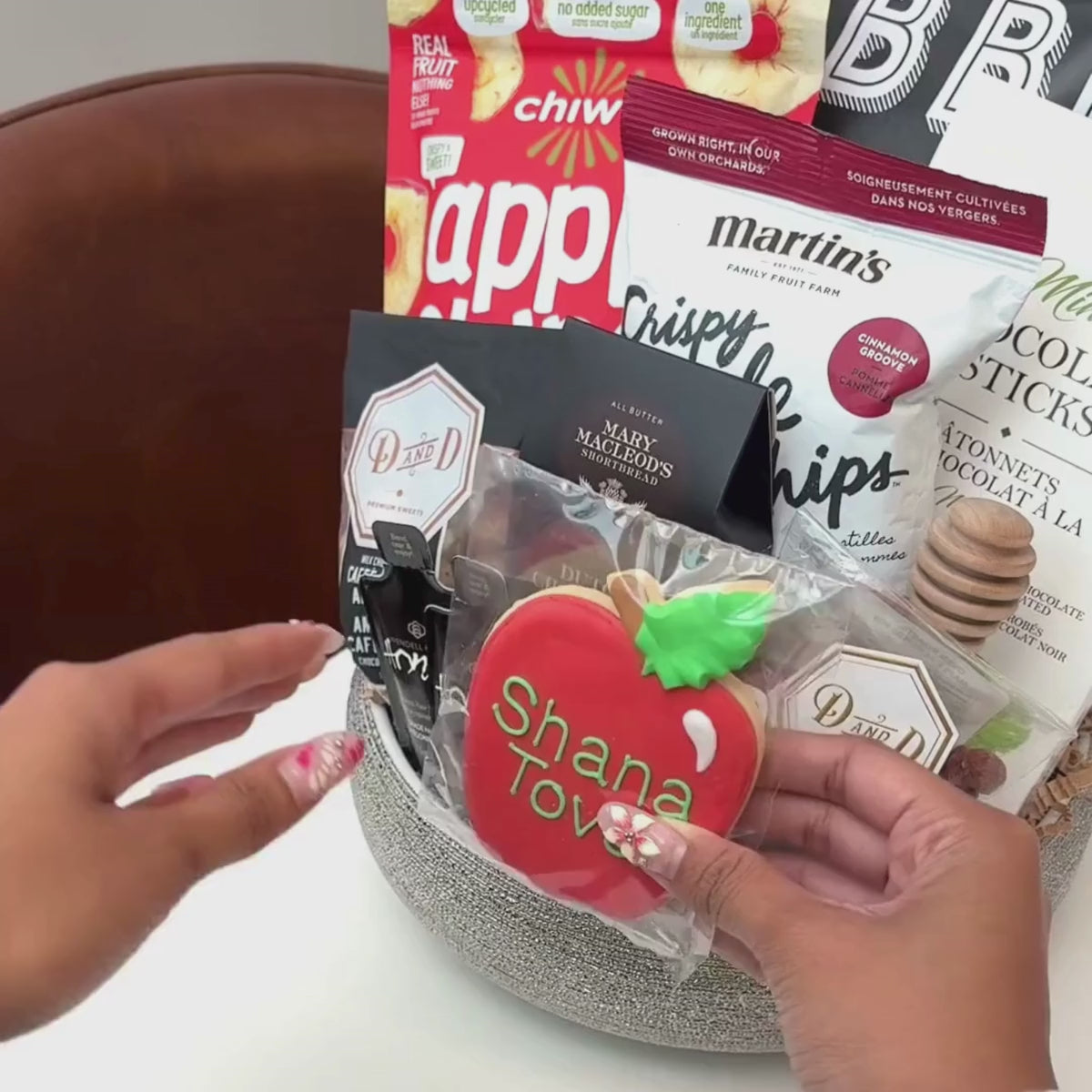 Hands unpacking an 11-piece Shana Tova gift basket, displaying a traditional, Hand-Painted Shana Tova Apple-Shaped Cookie and an array of festive indulgences.