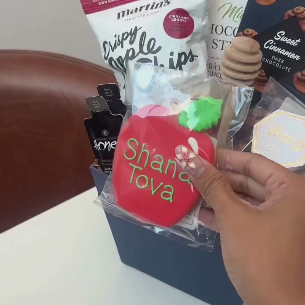 Hands unpacking an 8-piece Days of Awe gift basket, displaying a traditional, Hand-Painted Shana Tova Apple-Shaped Cookie, Honey Sachets and an array of festive indulgences.