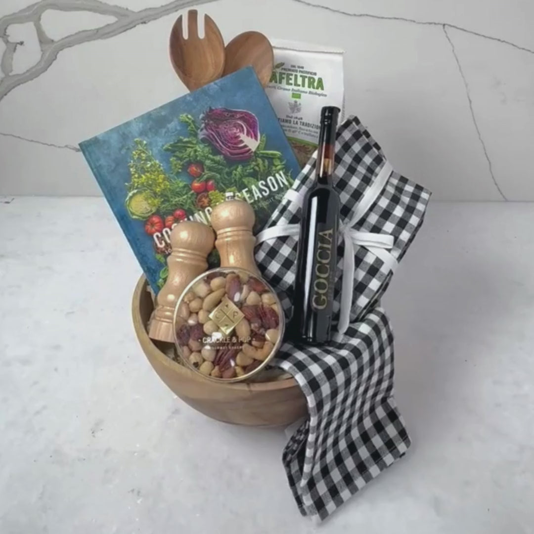 Hands unpacking a 9-piece Salad Bowl gift basket, displaying a bottle of Goccia 8-Year-Old Balsamic Vinegar, an Acacia Wood Spoon & Fork Set, along with an array of essential cooking items. 