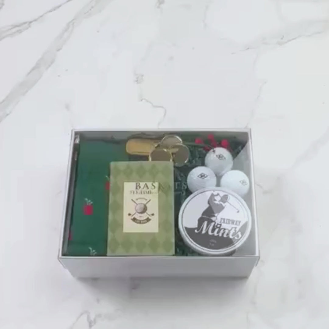 Hands unpacking a 9-piece Fore the Love of Golf gift box, displaying a Tee Time Golf notebook and an array of essential golf items.