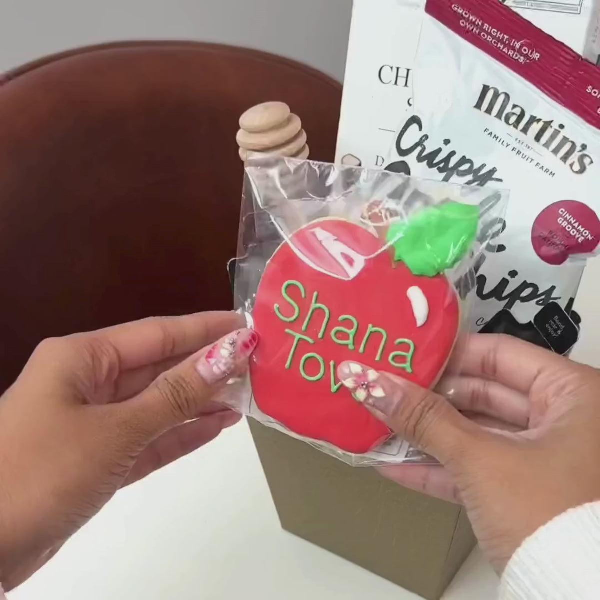 Hands unpacking the 6-piece Gilded Orchard gift basket, displaying a Hand-Painted Shana Tova Apple-Shaped Cookie, Honey Sachets, and an array of festive indulgences. 
