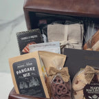 Hands unpacking a 19-piece Adelaide Gift Basket, displaying a Beige Cashmere Blanket and an array of Pancake Mix, Maple Syrup, Linen Napkins, and a Wooden Cutting Board.